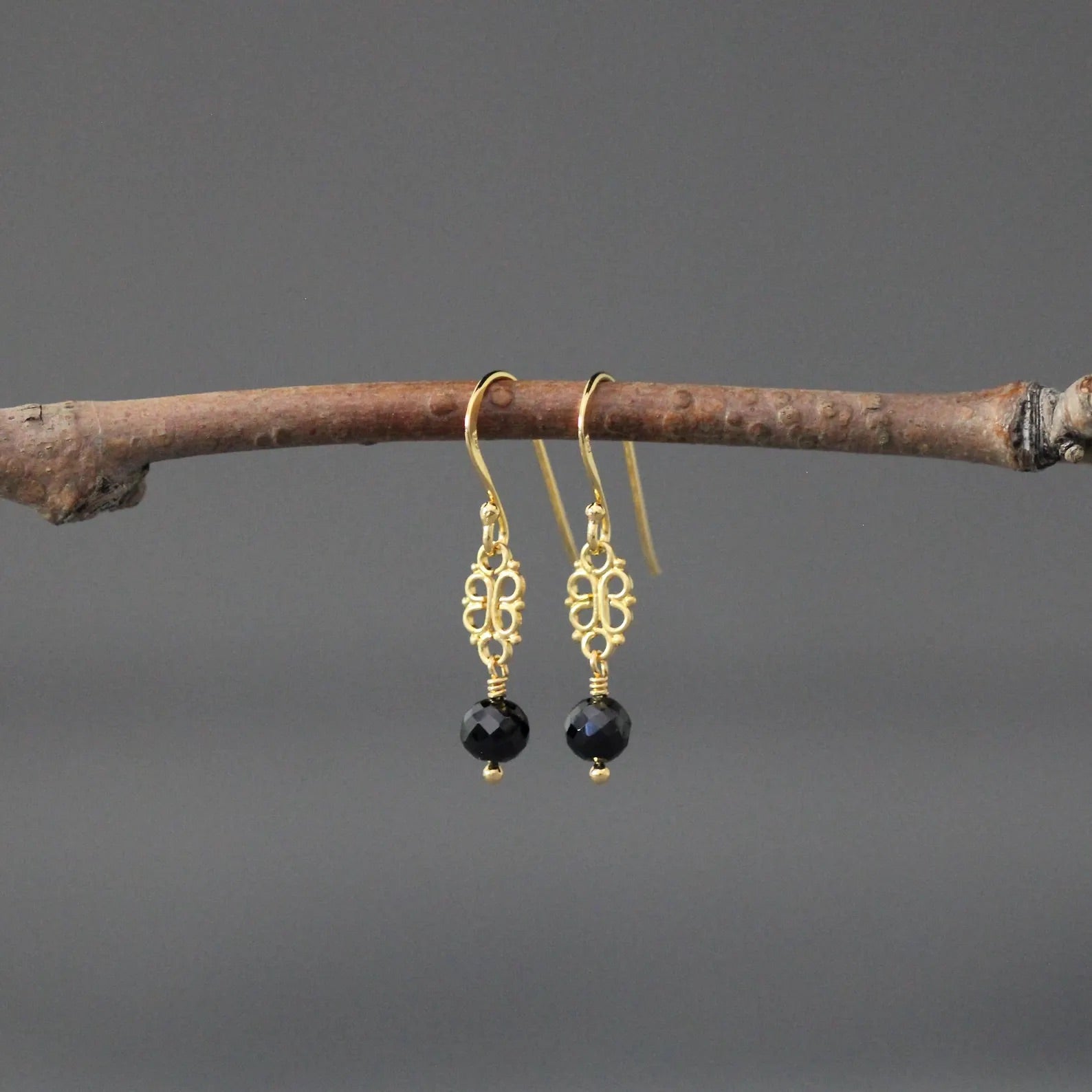 Gold earrings with black beads hanging on a branch against a gray background