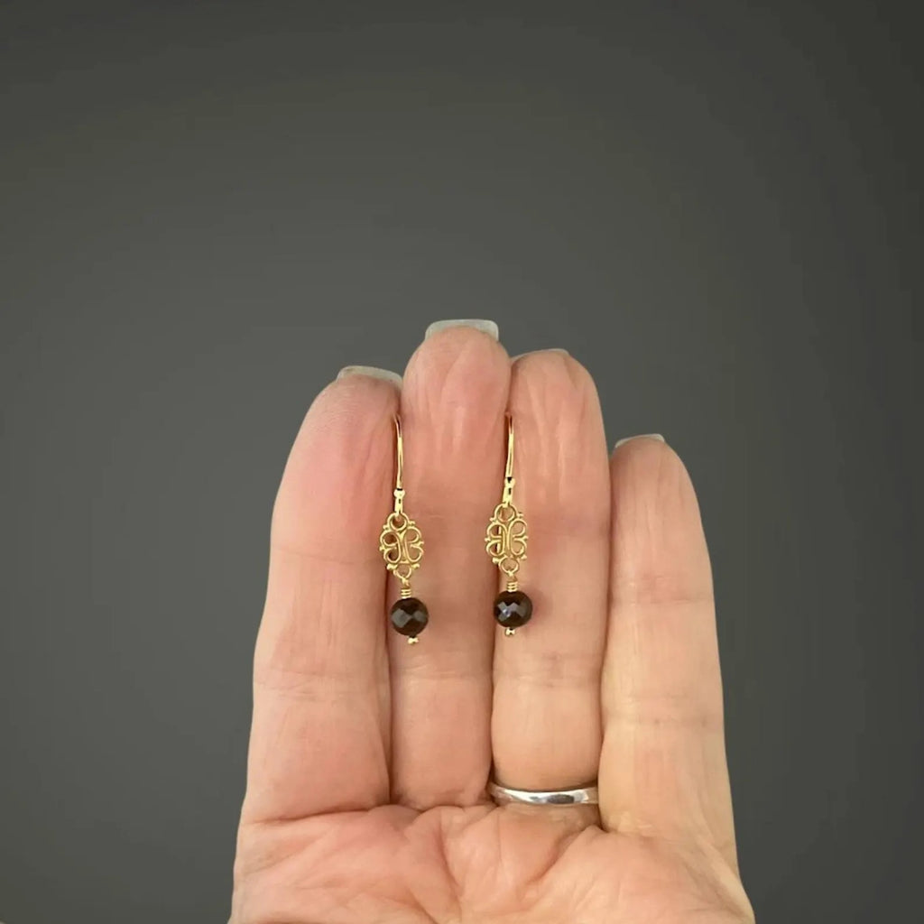 Gold earrings with dark gemstones held in a hand against a gray background