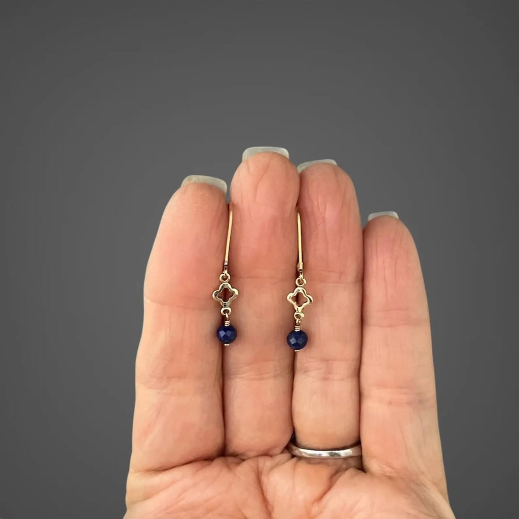 Gold hoop earrings with blue lapis teardrop and quatrefoil charms held in hand
