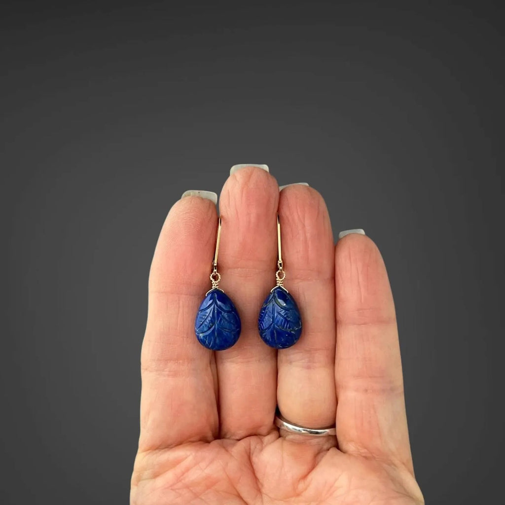 Blue carved lapis leaf teardrop earrings with gold leverback hooks, held in hand