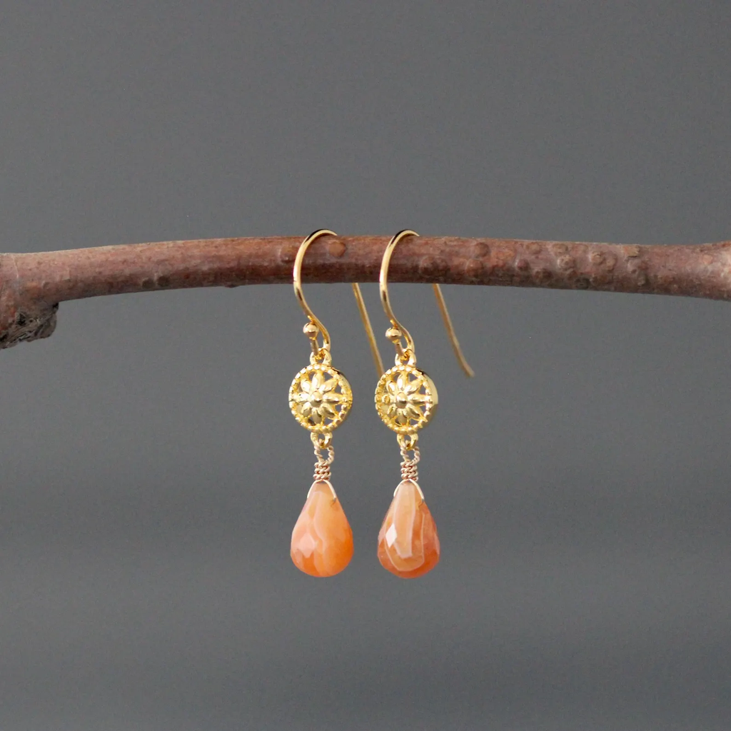 Gold earrings with orange teardrop stones hanging on a branch against a gray background