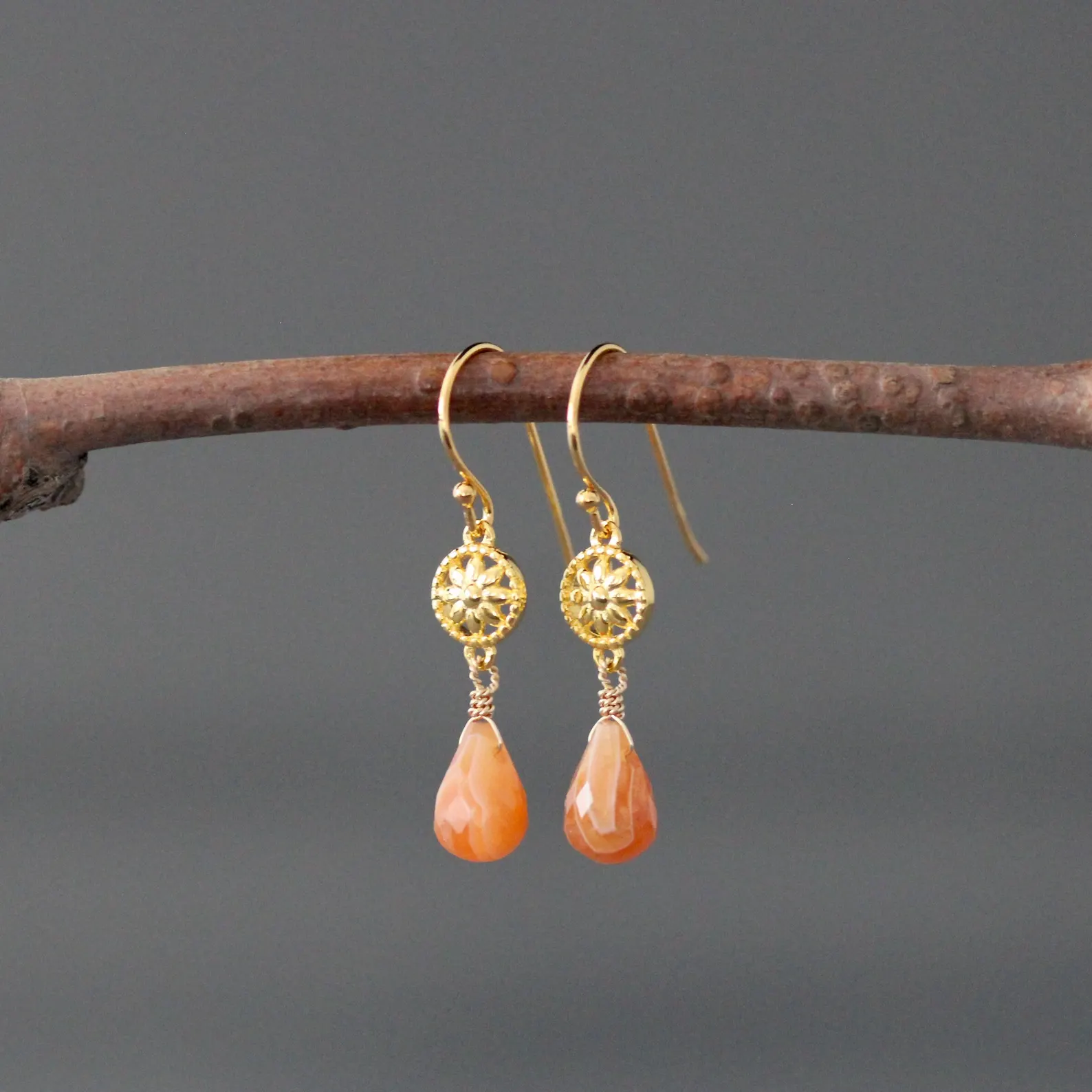 Gold earrings with orange teardrop stones hanging on a branch against a gray background
