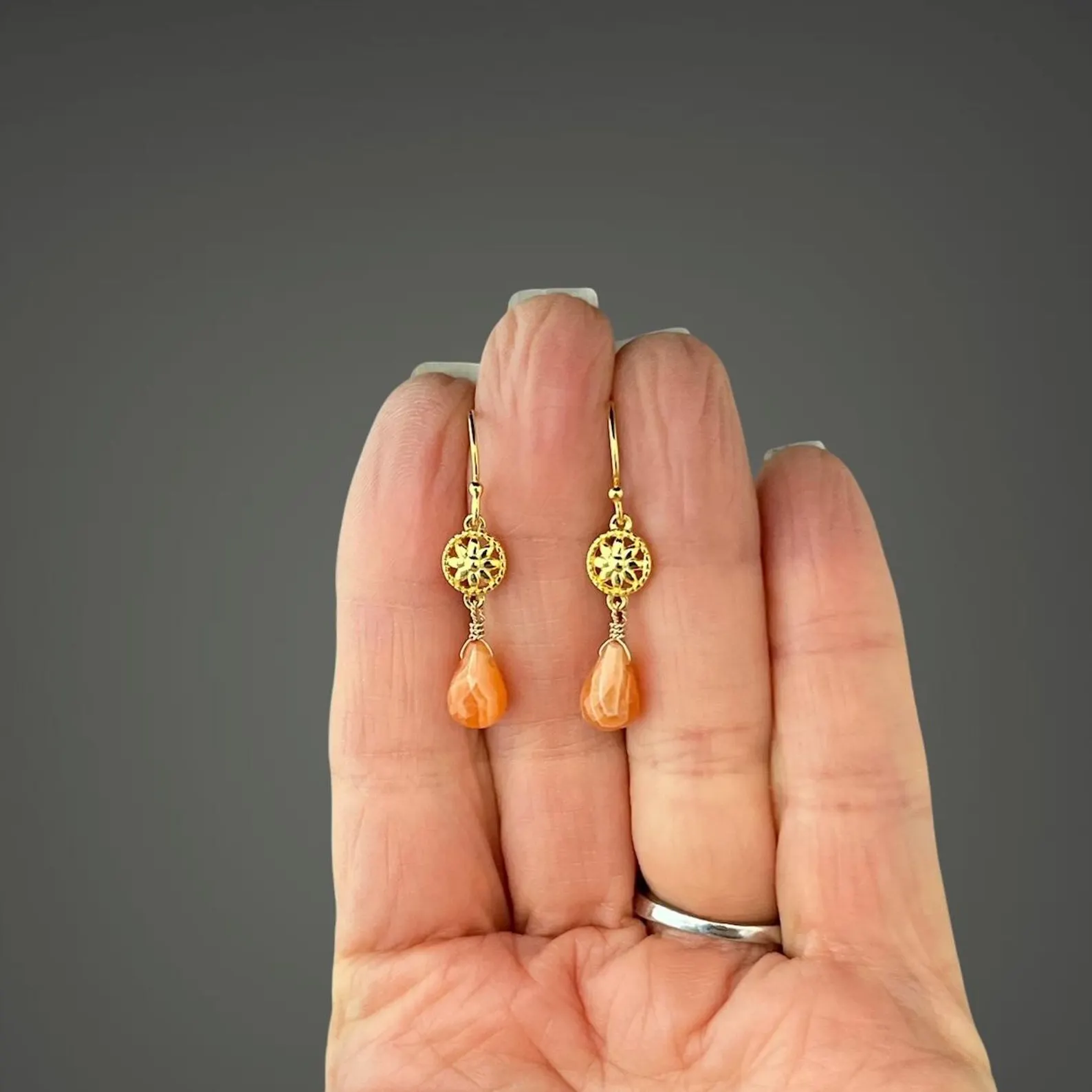 Gold earrings with orange beads held in a hand against a gray background