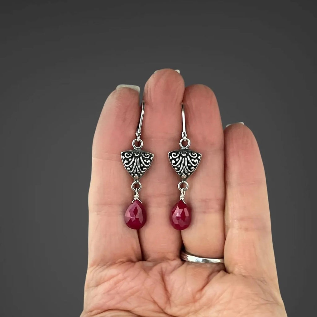 Silver earrings with ornate Bali-style tops and red ruby teardrop drops on hand