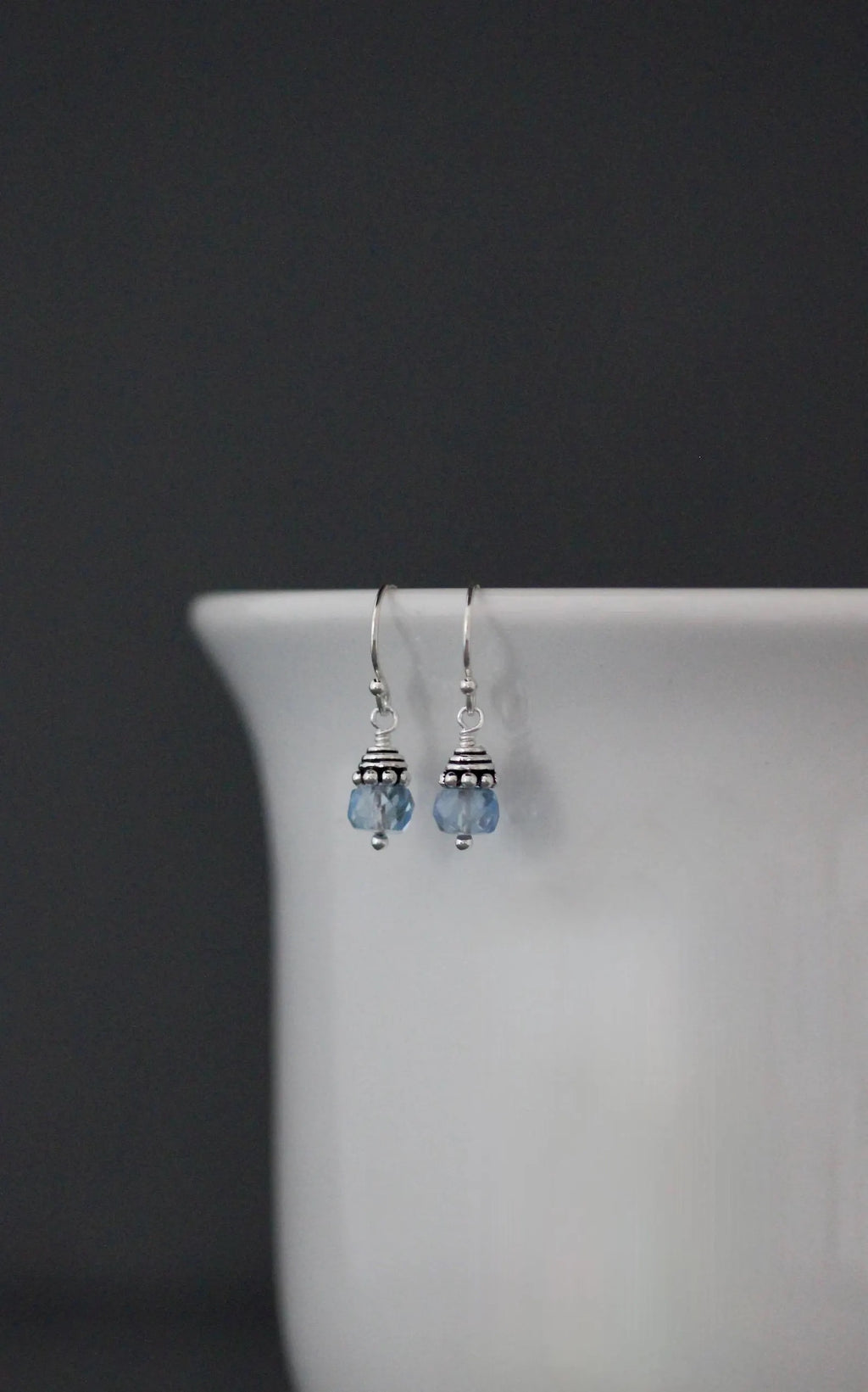 Pair of silver earrings with blue beads hanging on a white vase against a dark background