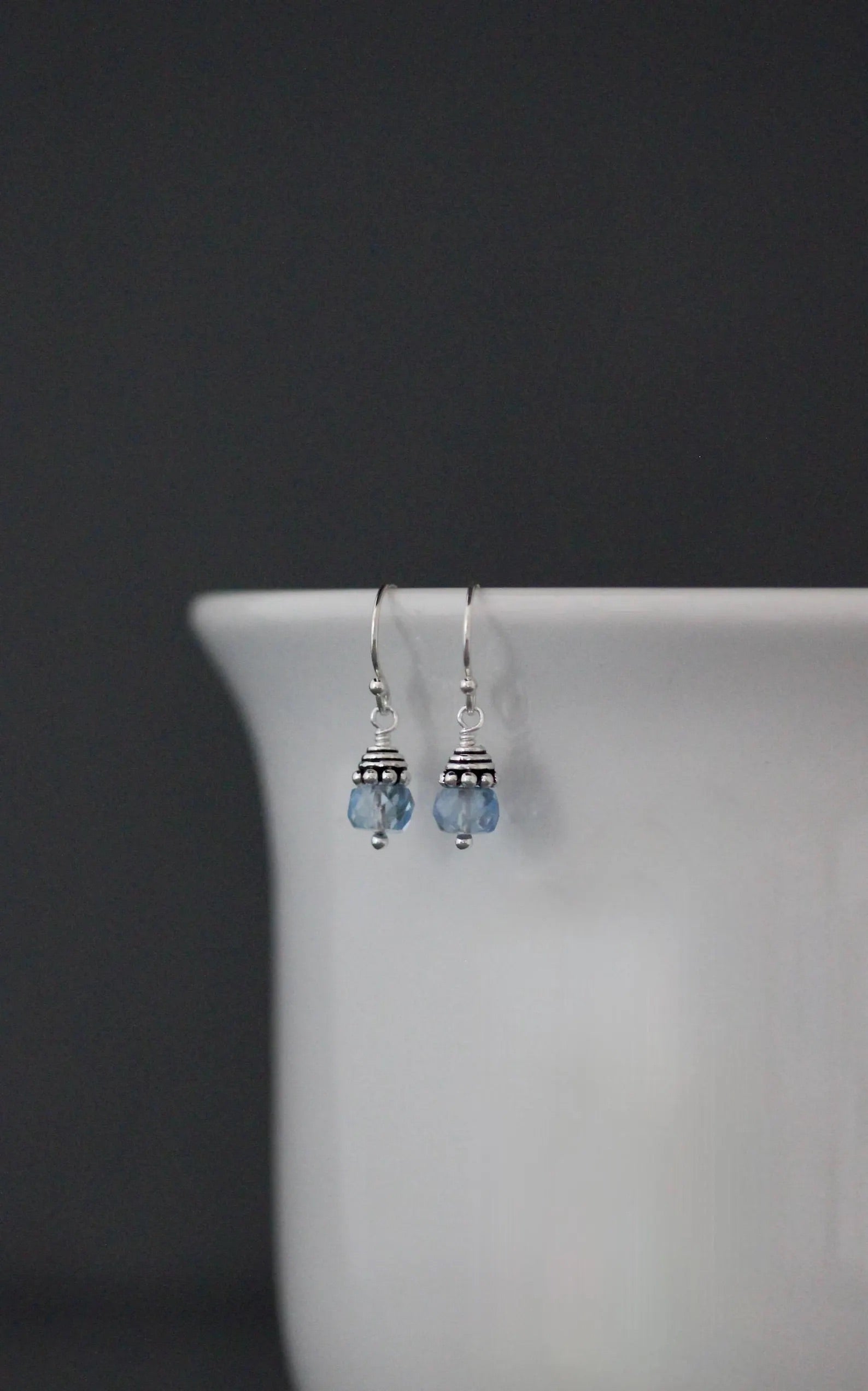 Pair of silver earrings with blue beads hanging on a white vase against a dark background